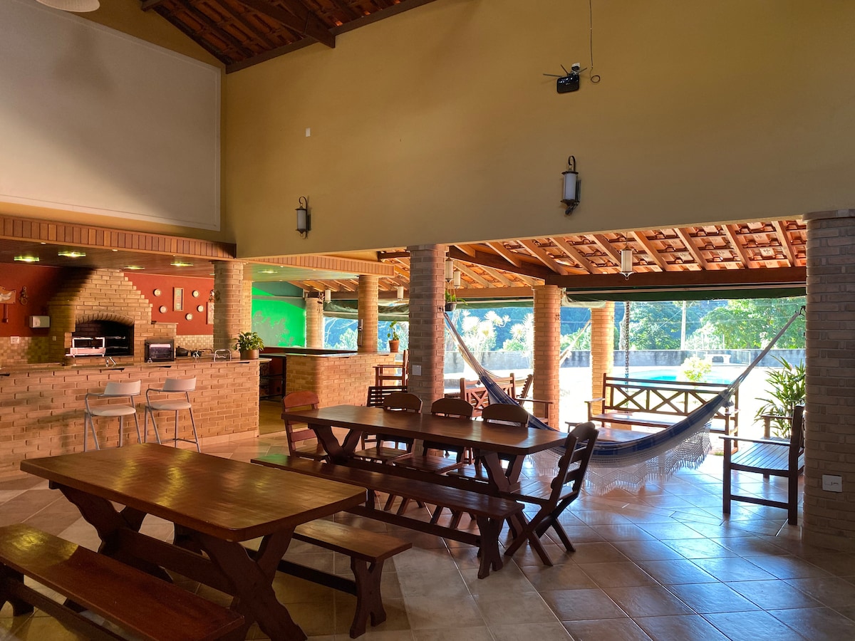 The spacious covered dining area features multiple wooden tables and a comfortable seating arrangement. A well-equipped kitchen is visible in the background, complemented by large windows that offer views of the outdoor space. Natural light illuminates the area, enhancing a relaxed ambiance.