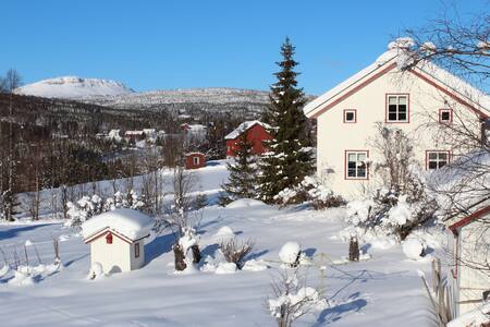 A winter landscape showcases a charming farmhouse with a sloped roof, surrounded by a thick blanket of snow. Sparse trees and snow-covered shrubs dot the landscape, while distant hills provide a scenic backdrop under a clear blue sky.
