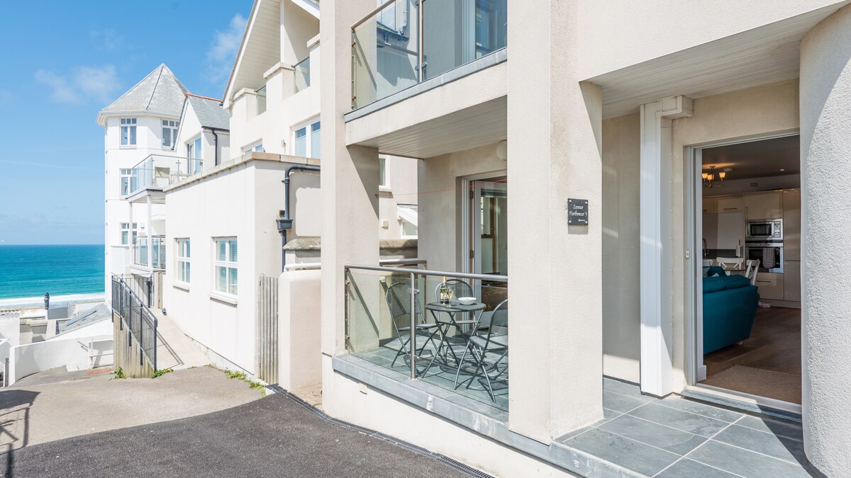 A coastal view is visible from the ground-floor balcony, which features a small outdoor seating area with a table and chairs. The adjacent entryway leads into the spacious living area, where large windows allow natural light to fill the space.