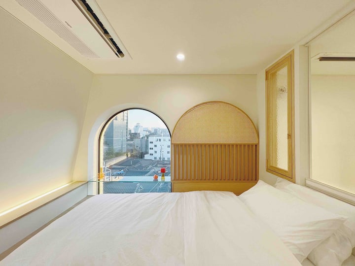 舒适的阁楼卧室，可欣赏传统韩屋屋顶的美景。 弧形窗户和藤条细节为这片宁静而明亮的空间增添了魅力。