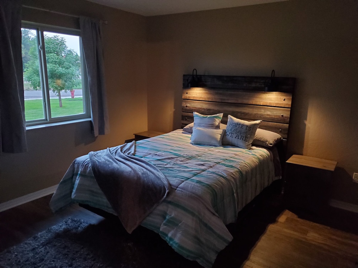 A welcoming bedroom is shown with a large bed featuring a wooden headboard. Soft bedding in light blue and white stripes is complemented by decorative pillows. Natural light filters through a window with sheer curtains, and warm wooden flooring is visible.