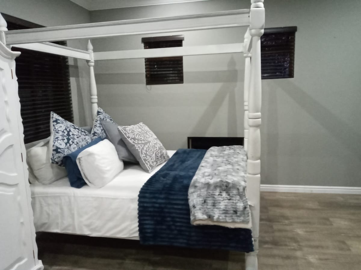 A four-poster bed is positioned against a muted wall, adorned with an arrangement of patterned and solid pillows. A textured throw drapes over the bed, contrasting with the light bedding. Two small windows, fitted with dark blinds, allow for filtered light in the room.