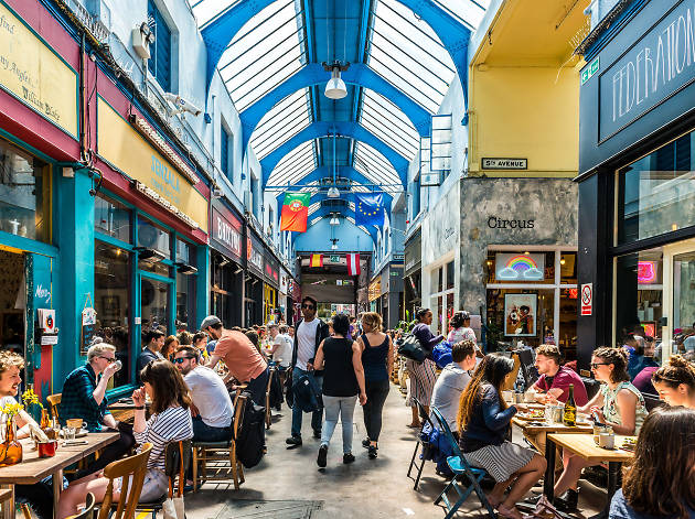 A lively marketplace is depicted, featuring colorful flags hanging from the ceiling. Tables are occupied by diners, with a mix of seating arrangements visible. Brightly painted walls and natural light streaming through skylights enhance the vibrant atmosphere.