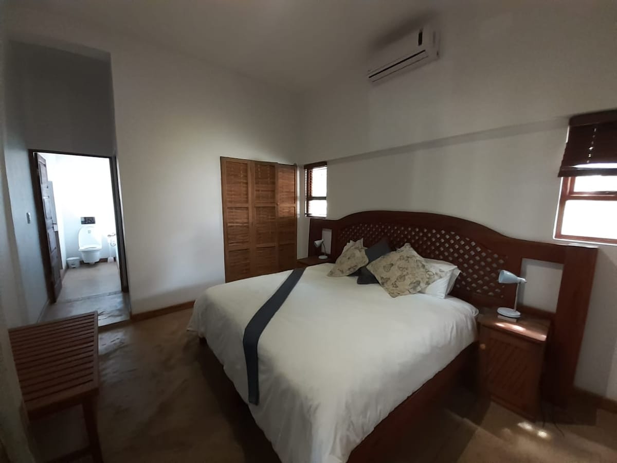 The airy bedroom features a spacious bed adorned with decorative pillows. Natural light enters through multiple windows, highlighting the wooden furnishings, including a bedside table and a slatted wooden chair. A doorway leads to an adjoining area, enhancing the room's openness.