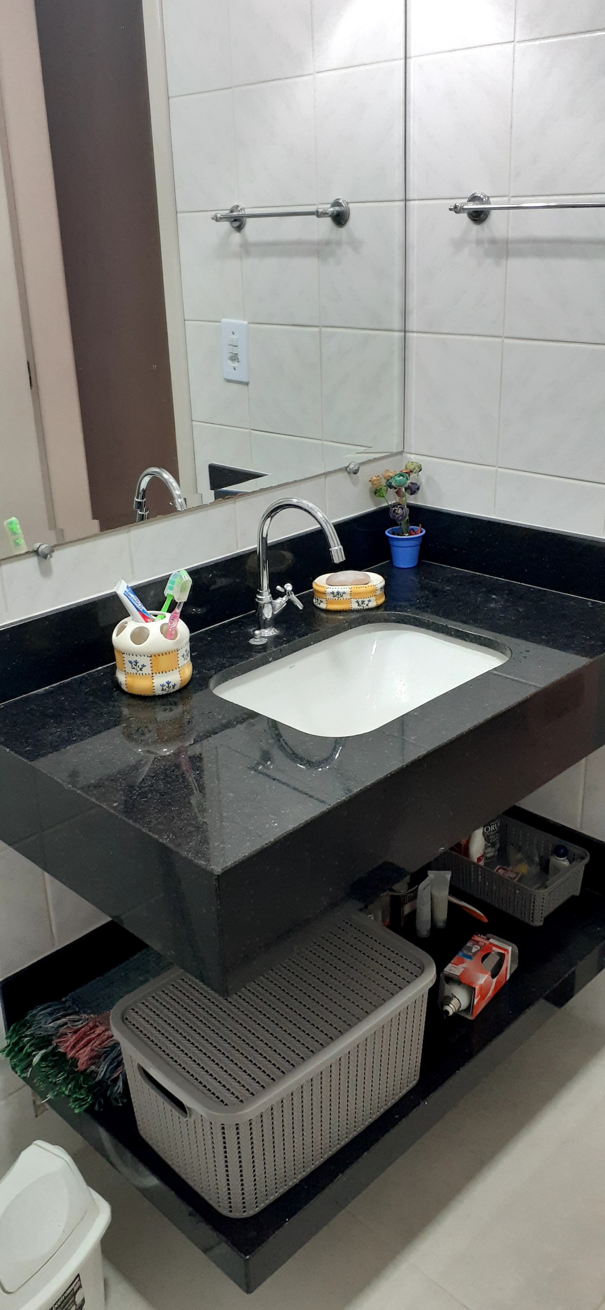 A sleek bathroom countertop made of dark granite showcases a rectangular sink and modern faucet. Storage containers and a woven basket are positioned underneath, while colorful decorative items are arranged on the countertop. The walls are tiled in light neutral tones.
