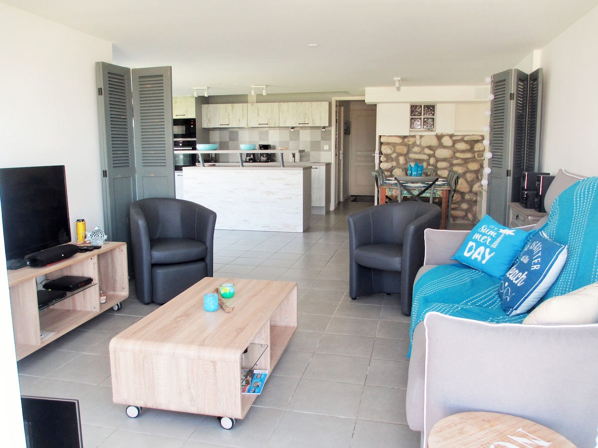 A spacious living area combines comfort and functionality. The modern kitchen, featuring light cabinetry, is visible in the background. A cozy seating arrangement includes a gray sofa and armchairs, while a wooden coffee table sits centrally, offering a relaxed space for guests to gather.