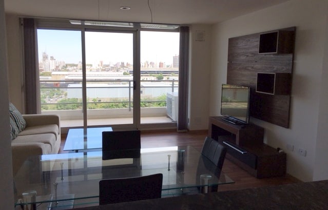A living area features a contemporary glass dining table surrounded by black chairs. A light-colored sofa is positioned nearby, offering views through large sliding glass doors that open to a balcony. The city skyline and river can be seen in the background.