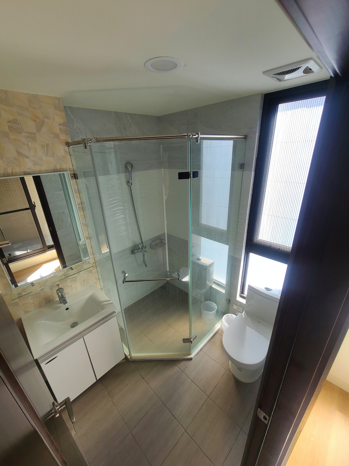 A modern bathroom features a glass-enclosed shower, a compact sink with storage beneath, and a toilet. Natural light filters through frosted windows, enhancing the clean and functional design.