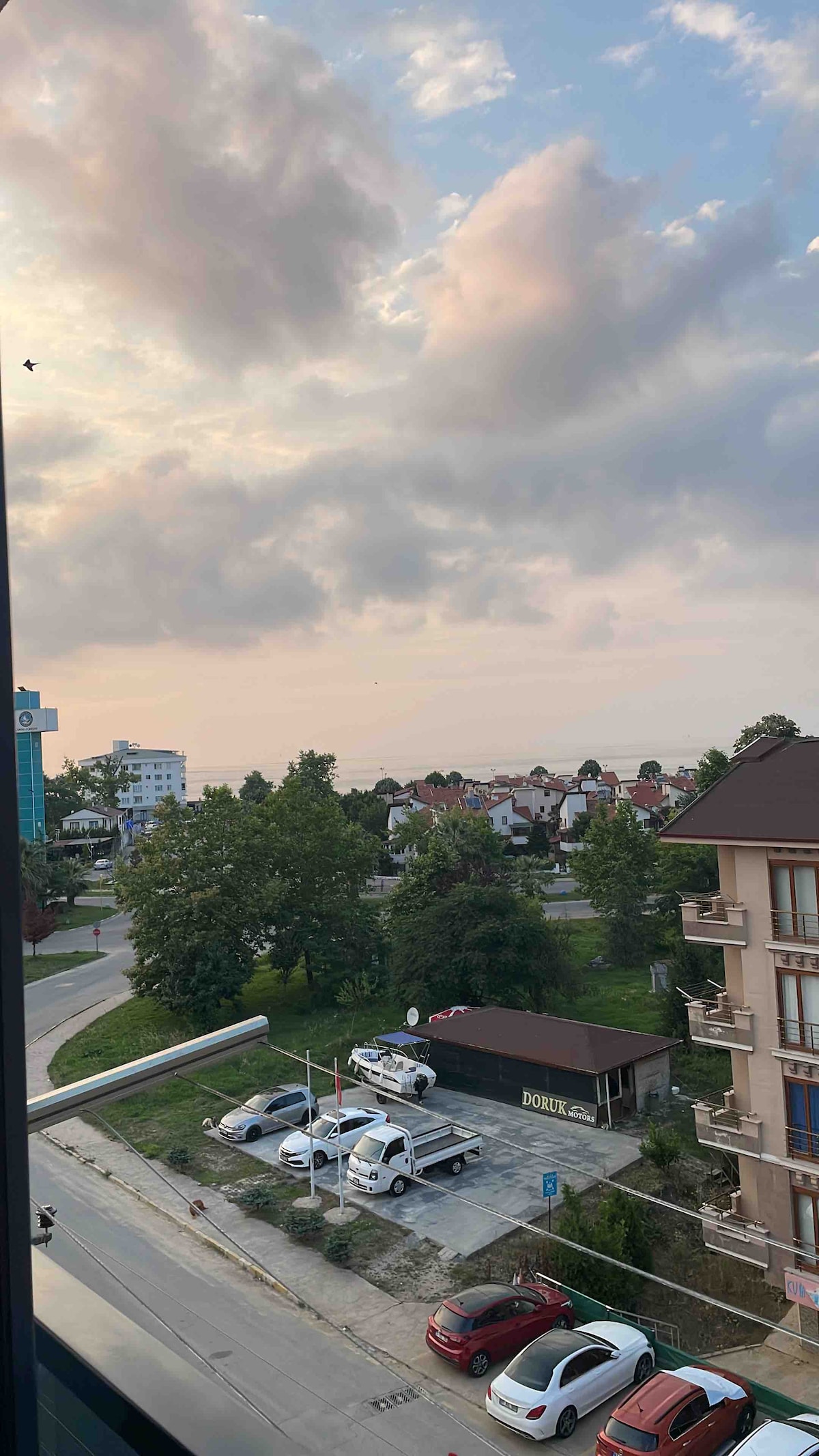 A view from a balcony showcases a green area with various trees and residential buildings in the distance. A road winds through the scene, leading to a parking area with vehicles. The sky is partly cloudy, suggesting a tranquil ambiance.