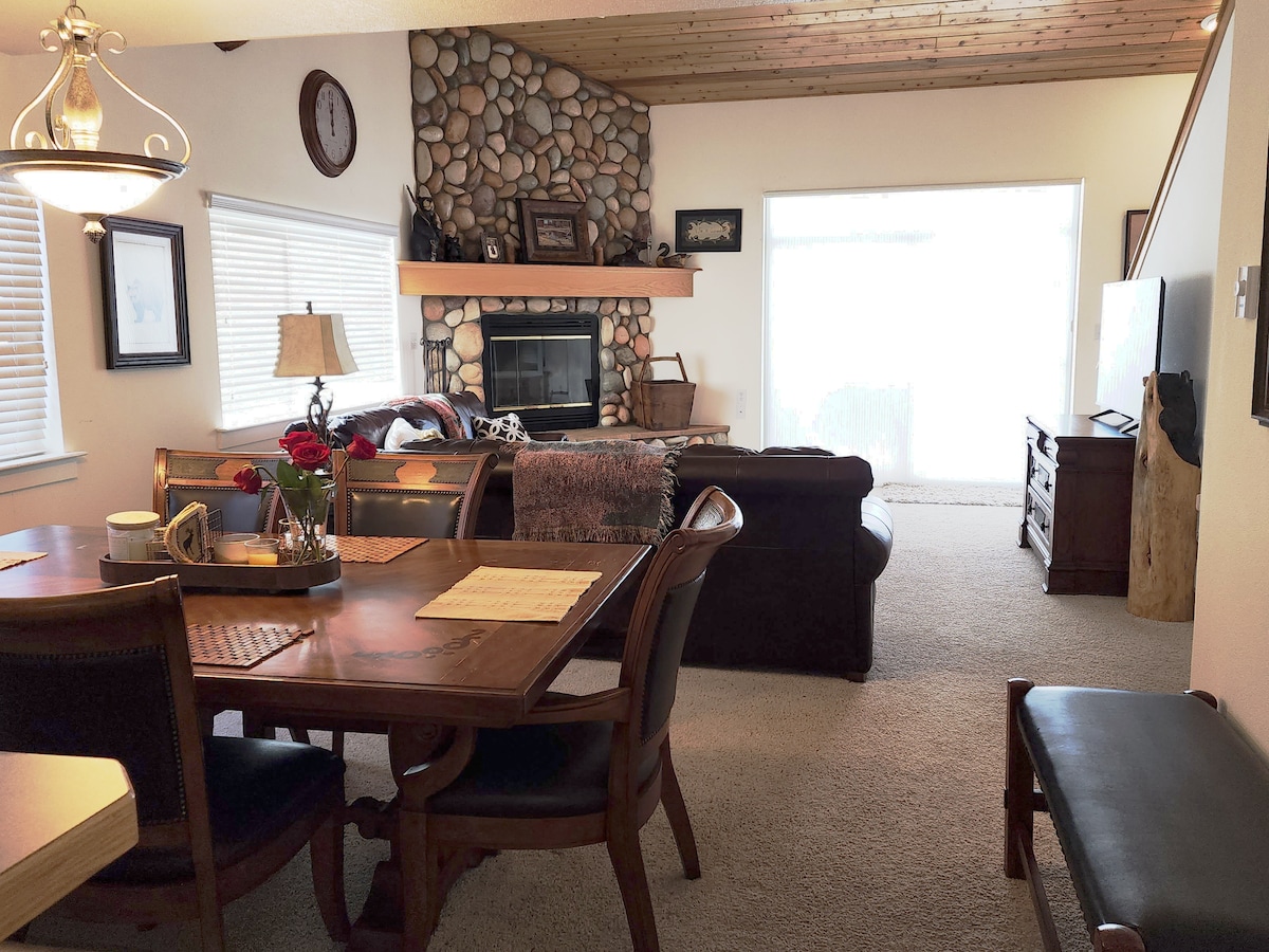 The living and dining area features a wooden table with seating for four, accompanied by a cozy couch and a stone fireplace. Natural light enters through the large windows, enhancing the spaciousness of the room. A console has been positioned near the window, providing additional storage.