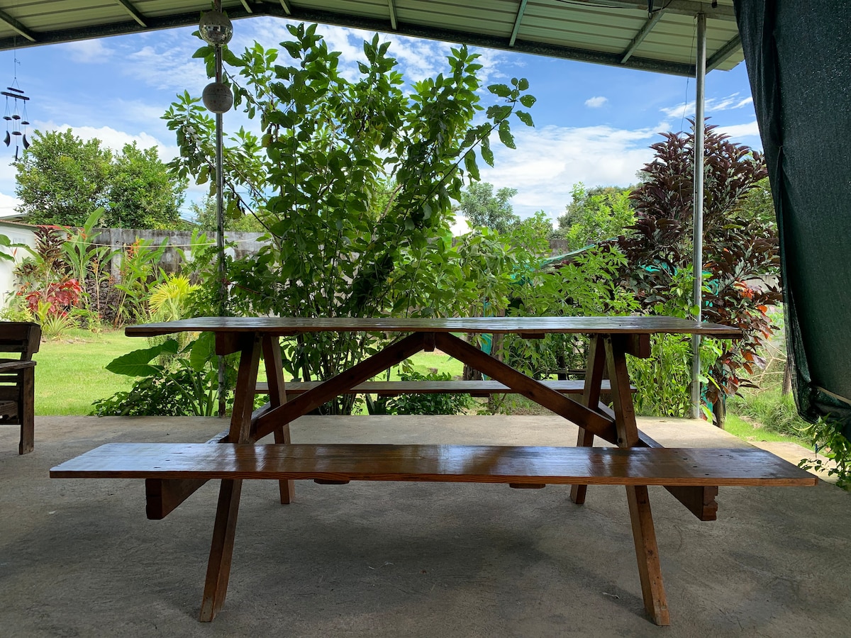 A rustic wooden picnic table is situated on a shaded terrace, complemented by matching benches. Lush green plants surround the area, providing a natural backdrop. The clear blue sky and scattered clouds are visible above, enhancing the tranquil atmosphere of the setting.