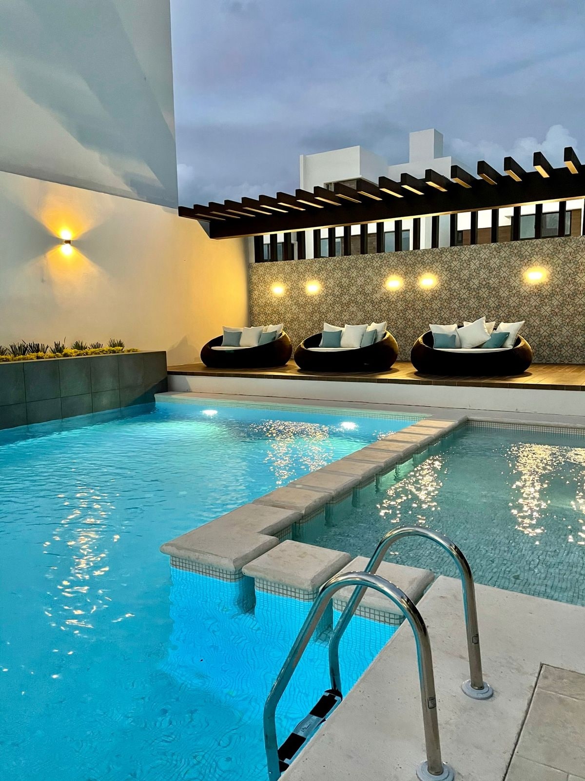A modern pool area features a shimmering turquoise pool, complemented by a cozy seating arrangement with black lounge chairs and bright cushions. Soft lighting accents the surrounding wall, creating a serene evening ambiance.