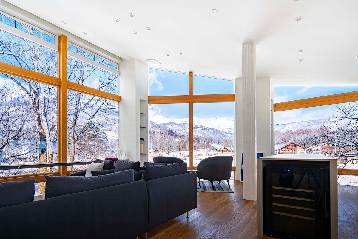 A spacious living area showcases large floor-to-ceiling windows which provide expansive views of the surrounding mountains. A modern sofa is positioned towards the windows, inviting relaxation. A sleek wine fridge is integrated into the kitchen area, complementing the refined decor of the chalet.