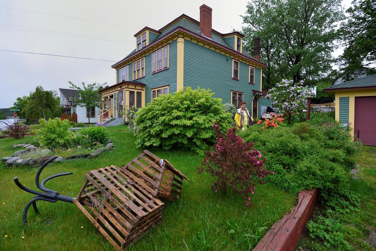 A charming multi-story house is surrounded by lush greenery and vibrant flowers. A wooden structure, possibly a bench or table, sits crookedly in the foreground. The house features colorful siding and multiple windows, providing a welcoming and inviting exterior.