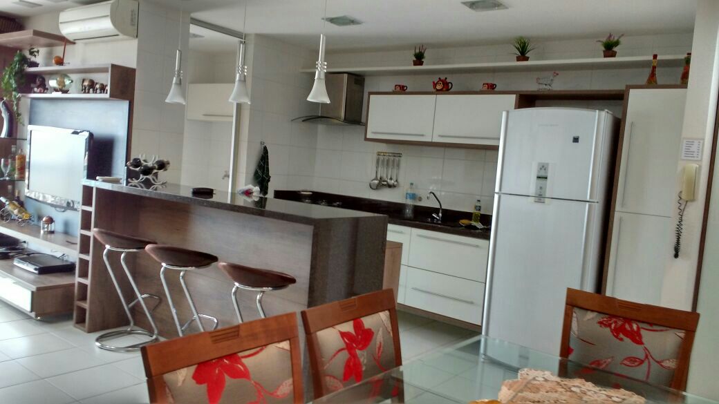 The open-concept kitchen and dining area features a modern design with a sleek countertop and seating for three at the bar. A refrigerator and various kitchen appliances are visible. The space is illuminated by pendant lights, with decorative elements arranged on shelves above.