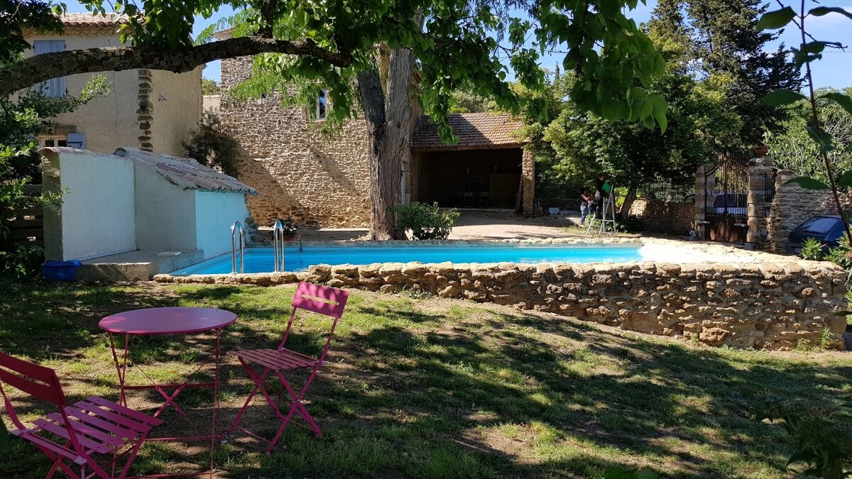 A serene outdoor area features a heated pool surrounded by stone walls. Two pink chairs and a table sit on the grassy lawn, offering a place to relax. Lush trees provide shade, and the rustic stone buildings add to the tranquil setting.