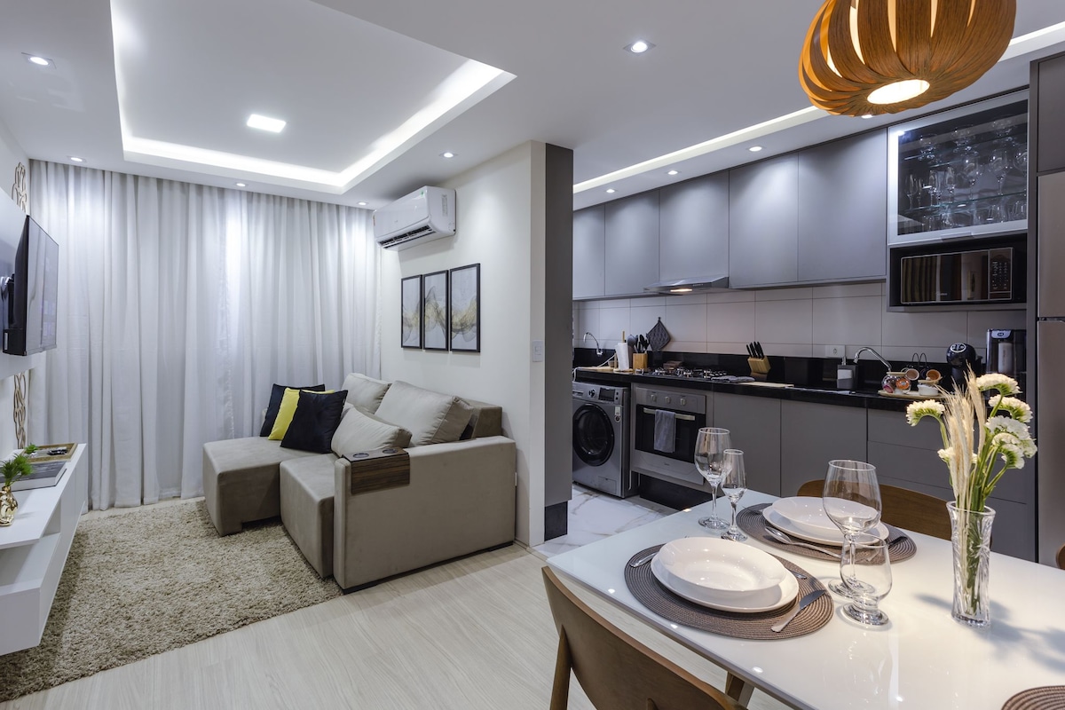 A modern living area features a sectional sofa positioned near a wall-mounted television. The adjacent kitchen is equipped with stainless steel appliances and neatly organized utensils. A dining table is set with glassware and plates, and soft lighting illuminates the space, complemented by decorative artwork.
