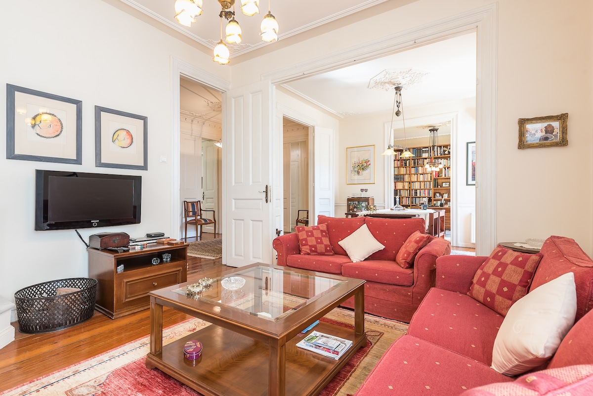 A living room features two red sofas arranged around a wooden coffee table, enhancing comfort. A flat-screen TV is mounted on the wall opposite the seating area, while artworks adorn the walls, contributing to a cozy atmosphere. Natural light fills the space through large doors.
