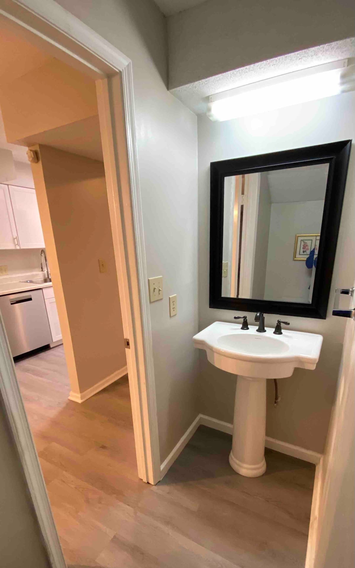 A compact half bath features a pedestal sink with modern faucets, complemented by a large rectangular mirror framed in black. Light-colored walls enhance the room's brightness, and the laminate flooring extends into the visible hallway, connecting to the adjacent kitchen area.