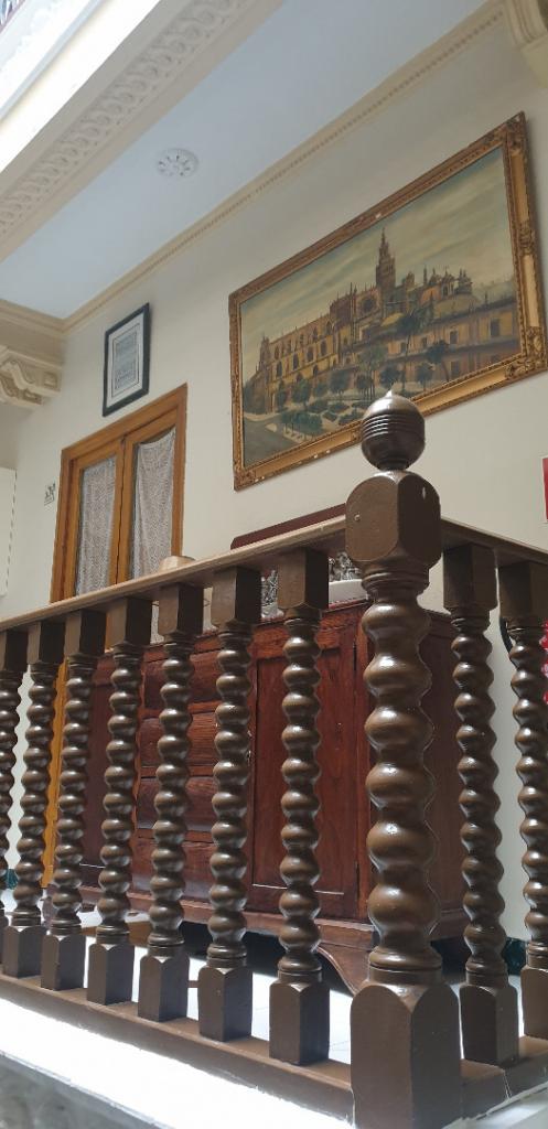 A decorative wooden railing with a spiral design is prominently featured in the foreground. A large painting depicting a historic structure is displayed on the wall above a wooden cabinet. Natural light illuminates the area, enhancing the traditional atmosphere.