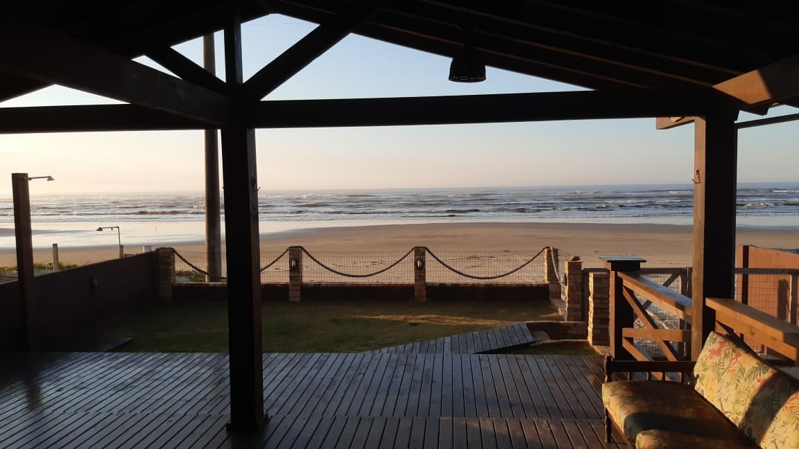 The image captures a beachfront view from a shaded wooden deck, showcasing rolling waves meeting the sandy shore. A rustic wooden beam structure frames the scene, while comfortable seating invites relaxation. The early morning light gently illuminates the landscape, enhancing the coastal atmosphere.
