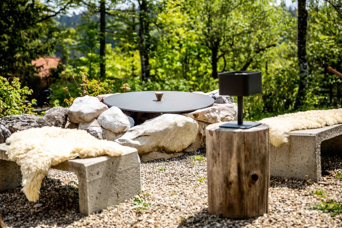 An outdoor seating area features two stone benches with soft cushions, arranged around a circular fire pit made of natural stones. A rustic wooden stool stands beside the fire pit, set against a backdrop of lush greenery and trees.