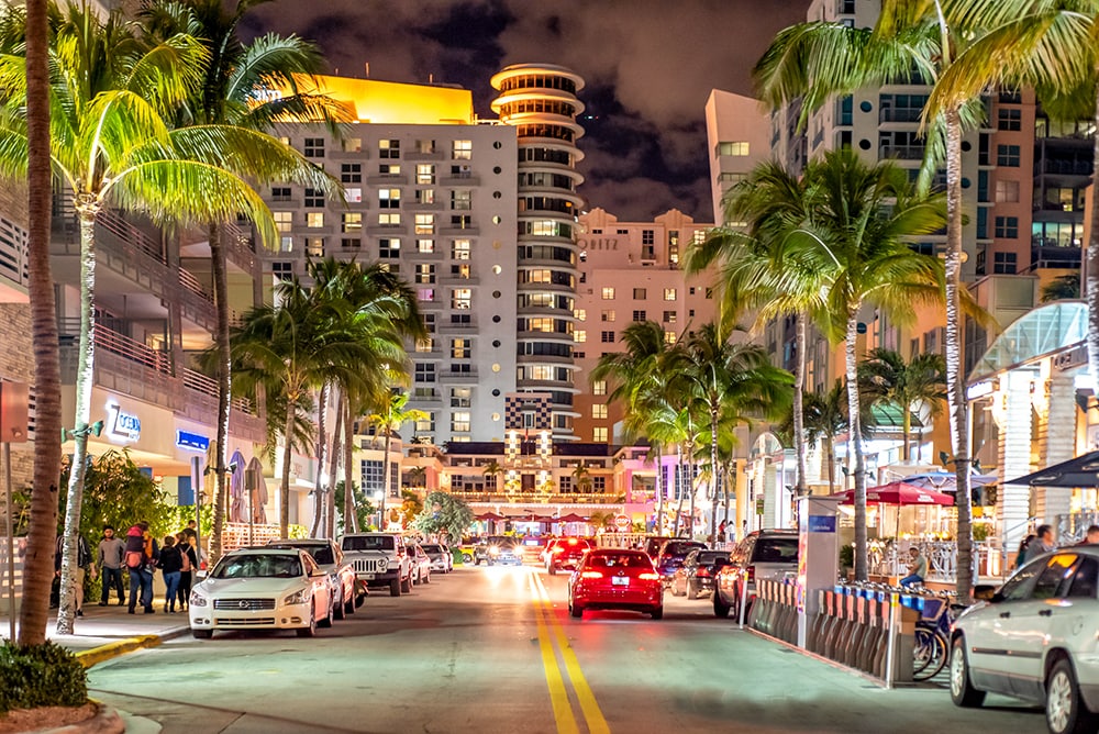 A lively street scene at night features illuminated buildings and palm trees lining the road. Cars are parked along the street, and colorful outdoor seating is visible in front of restaurants. Lights reflect off the buildings, creating a vibrant atmosphere in South Beach.