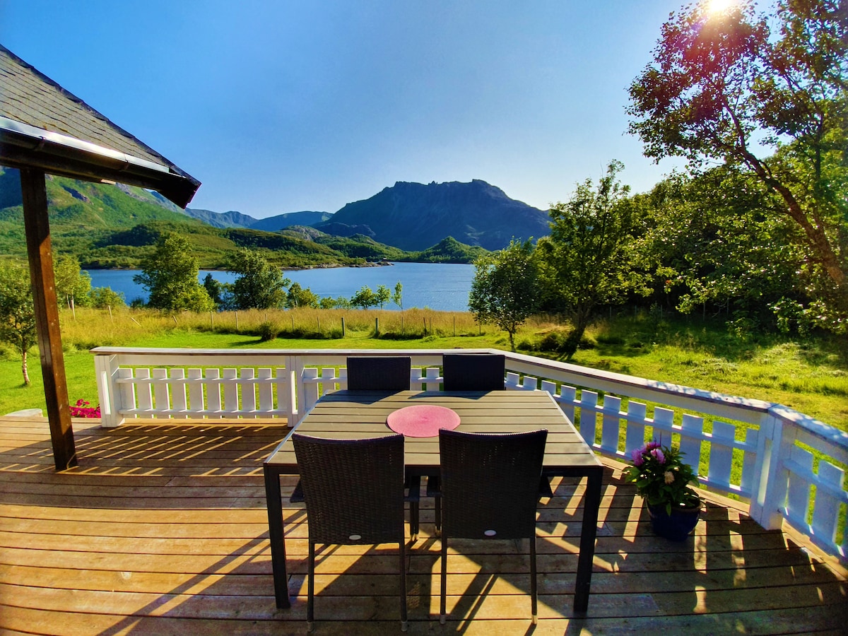 A spacious deck is seen overlooking a calm body of water, framed by lush greenery and mountains in the distance. A set of four dark wicker chairs surrounds a rectangular table, enhanced by a central pink placemat. Sunlight casts a warm glow across the scene.