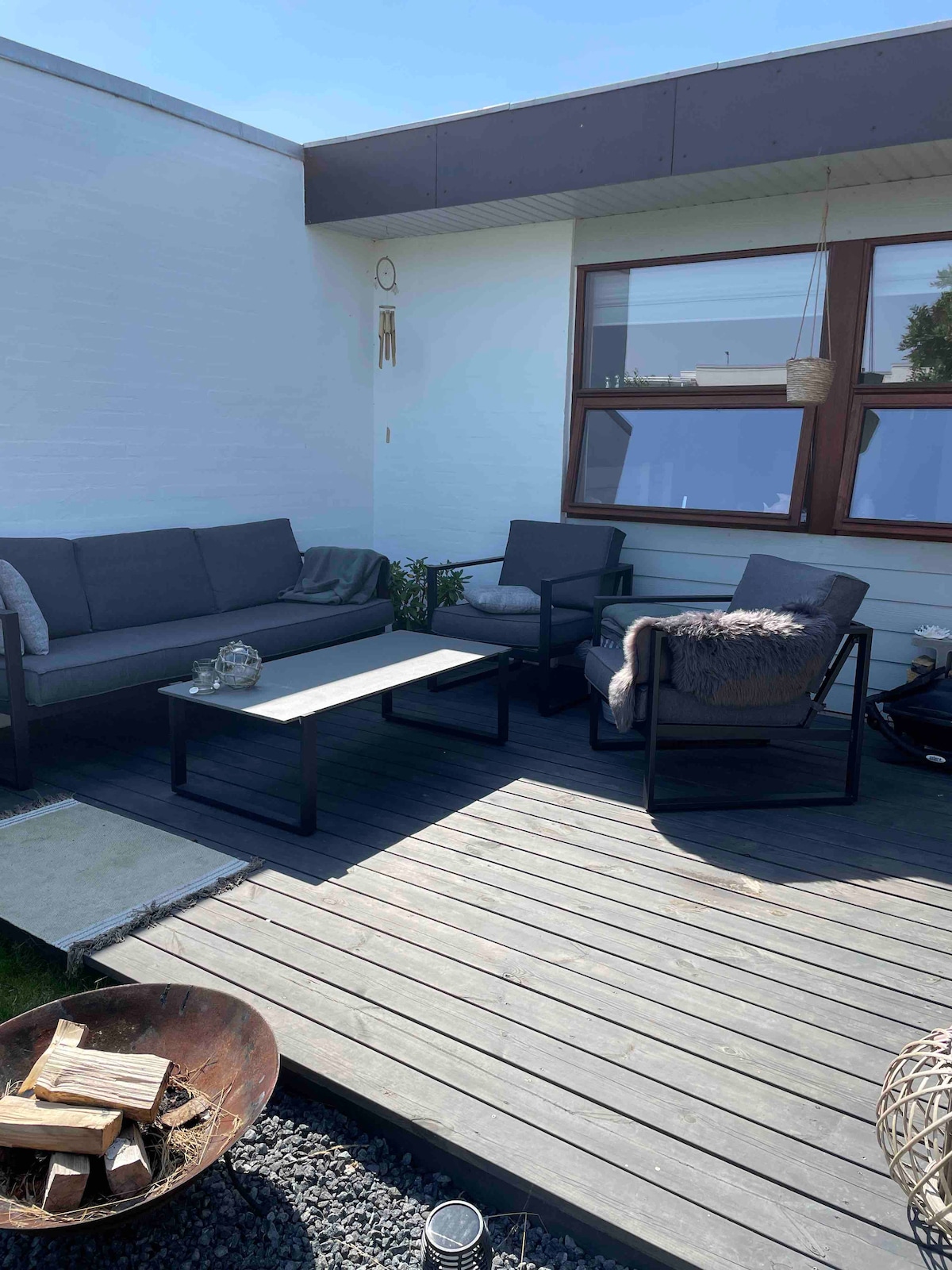 An outdoor seating area features a modern gray sofa and two matching chairs positioned around a rectangular table. Wooden decking provides a warm surface, while a small fire pit and decorative elements are placed nearby, creating a cozy space for relaxation.
