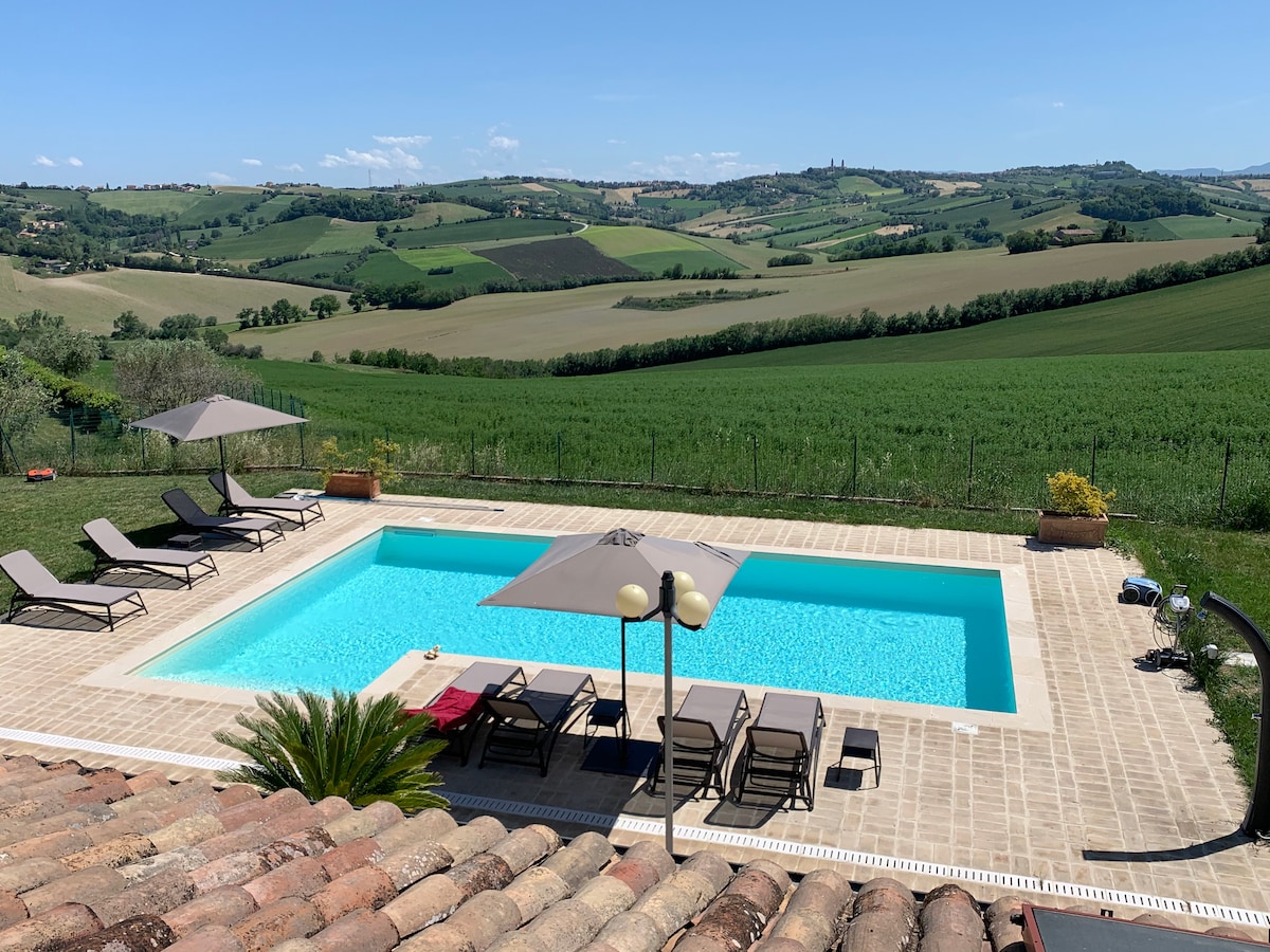 A serene outdoor swimming pool is surrounded by a paved area featuring multiple lounge chairs and umbrellas. Lush green hills and fields create a picturesque backdrop, while the clear blue sky adds to the tranquil setting.