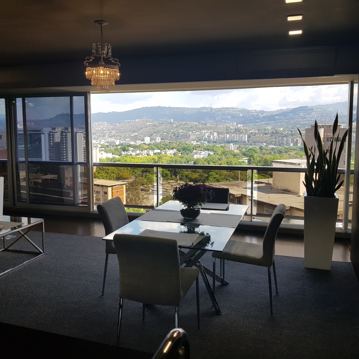 A dining area is visible, featuring a glass table surrounded by four upholstered chairs. A large window offers a panoramic view of lush hills and the city skyline. Natural light floods the space, accentuating the warm tones of the decor.
