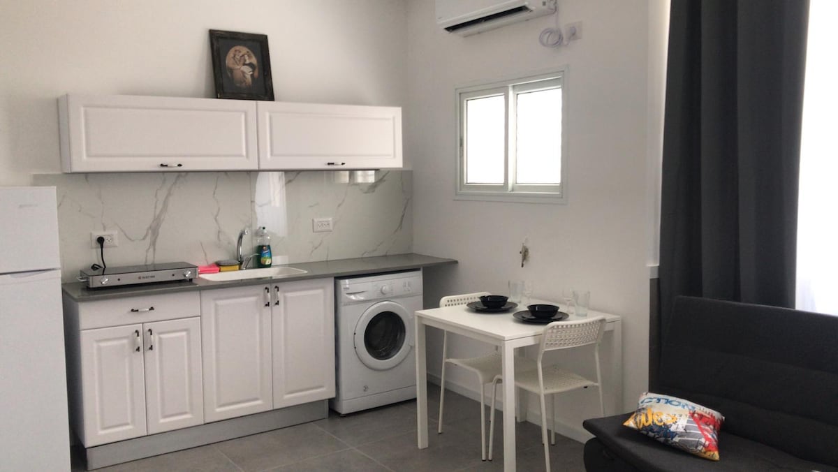 A modern kitchen area features white cabinetry and gray countertops. A washing machine and electric stovetop are included. A small dining table with two chairs is set against a wall, with light streaming in through a window adorned with light-colored curtains.