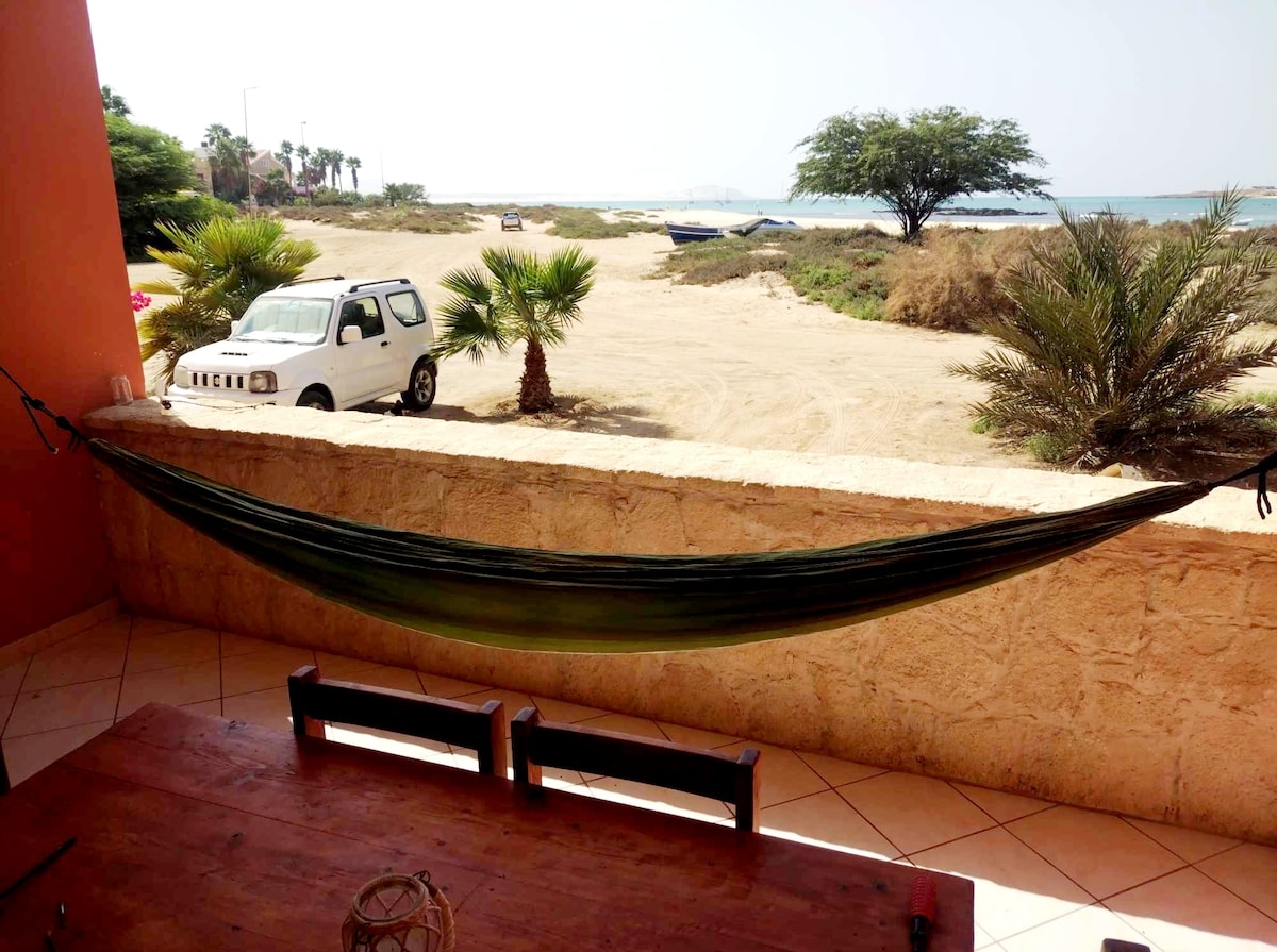 A green hammock is suspended from the terrace railing, providing a relaxing spot overlooking the outdoor landscape. Palm trees dot the sandy area, leading toward a distant view of the ocean. A simple table with chairs is positioned on the terrace, inviting outdoor gatherings.
