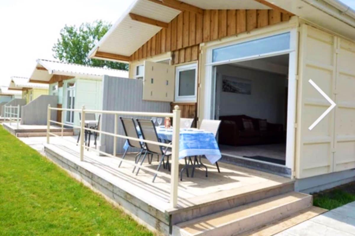 A spacious terrace is visible, featuring a dining table set with chairs. The lodge's sliding glass door opens to the interior, where a comfortable seating area is indicated. The building's wood and white design harmonizes with the green grassy surroundings.