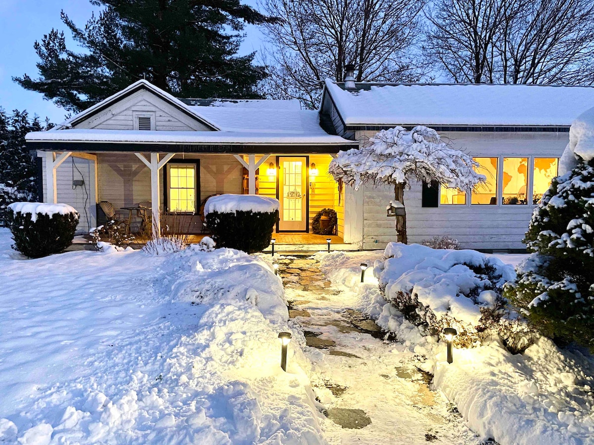 A welcoming farmhouse exterior is visible, surrounded by snow-covered landscaping. Warm light spills from the front door, highlighting the inviting porch area. Pathway lights illuminate the pathway leading to the entrance, creating a charming contrast with the winter scenery.