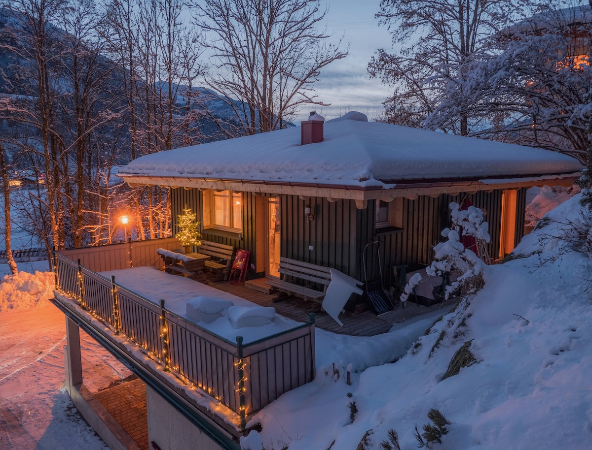 A modern wooden chalet is nestled in a snowy landscape, with a covered terrace adorned with warm lights. The structure features large windows that reflect the surrounding trees and the evening sky, creating an inviting winter scene.