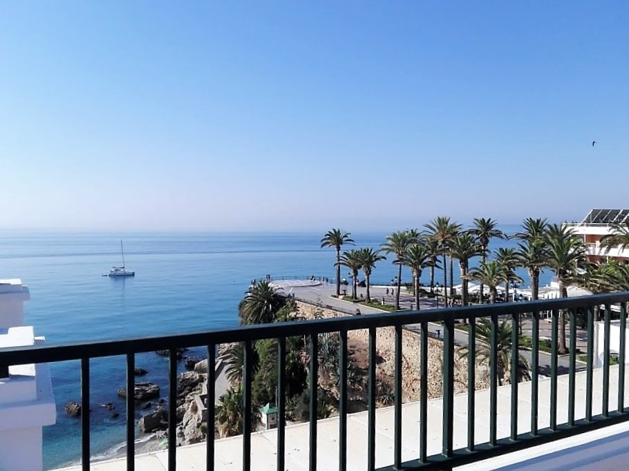 A panoramic view captures the serene ocean against a clear blue sky, framed by palm trees. The tranquil waters reflect the sunlight, with a small boat visible in the distance. A railing provides a perspective over the coastline and nearby promenade.
