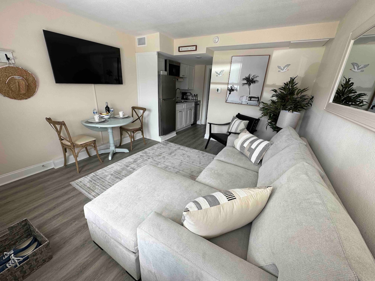 A modern living area is shown, featuring a light gray sectional sofa with decorative pillows. A small dining table for two is positioned nearby, alongside a wall-mounted flat-screen TV. The kitchen area is visible in the background, and potted greenery adds a touch of warmth.