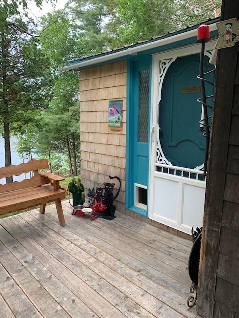 A charming entrance to the cottage is highlighted by a wooden bench and whimsical decor. The door features intricate designs, and colorful artwork is displayed nearby. Natural surroundings create a serene setting, with hints of the river visible through the trees.