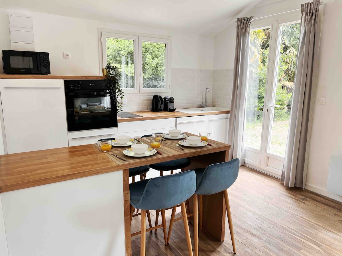 A bright kitchen features a wooden dining table set with plates and drinks. Modern appliances, including a microwave and oven, are present, alongside a sink and coffee maker. Large windows offer a view of the outdoors, complemented by light-colored cabinetry and stylish bar stools.