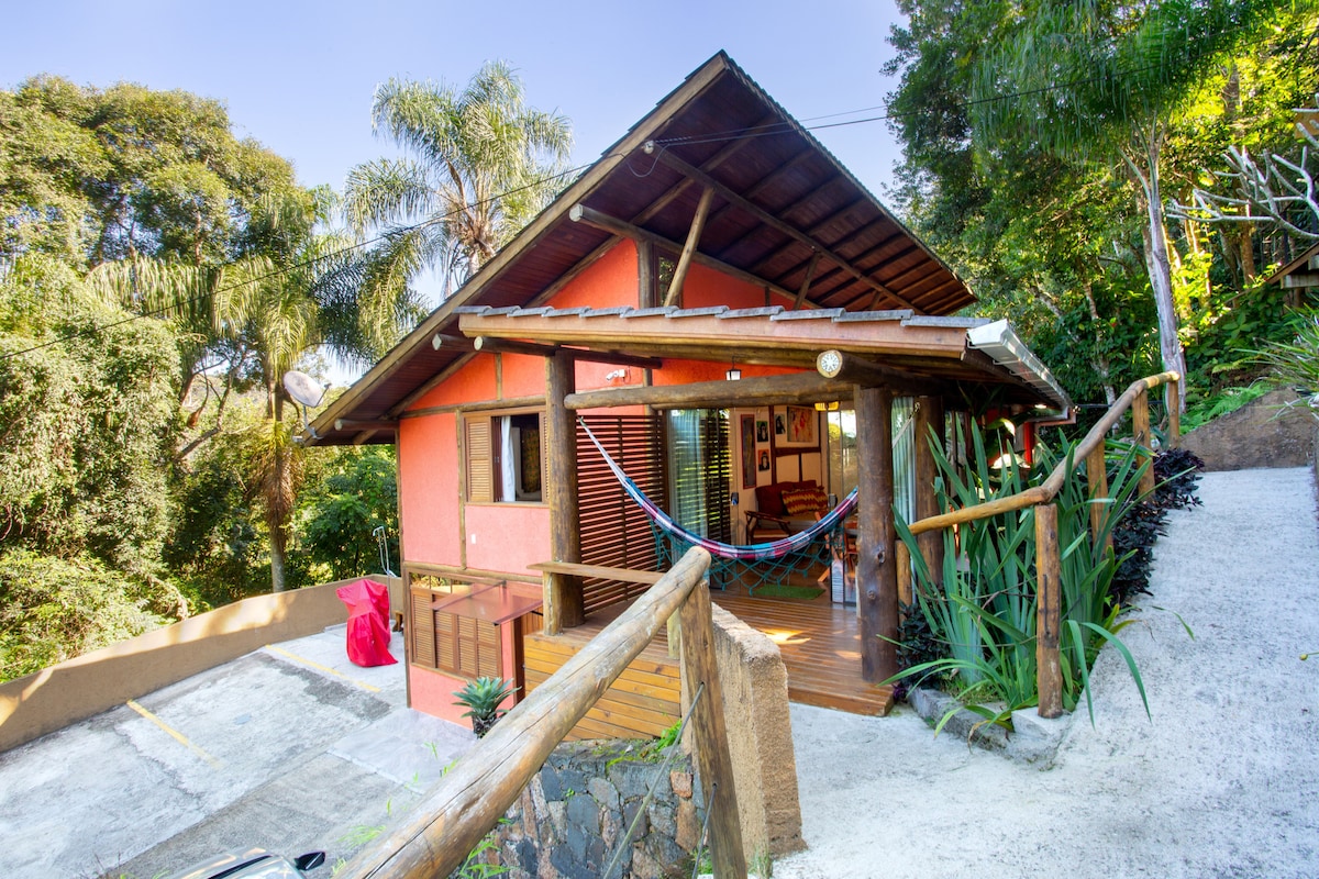 The exterior of a charming cabin is surrounded by lush greenery, featuring a wooden deck with a hammock and a rustic railing. Natural stone steps lead to the entrance, showcasing vibrant orange walls and a thatched roof blending with the tranquil environment.