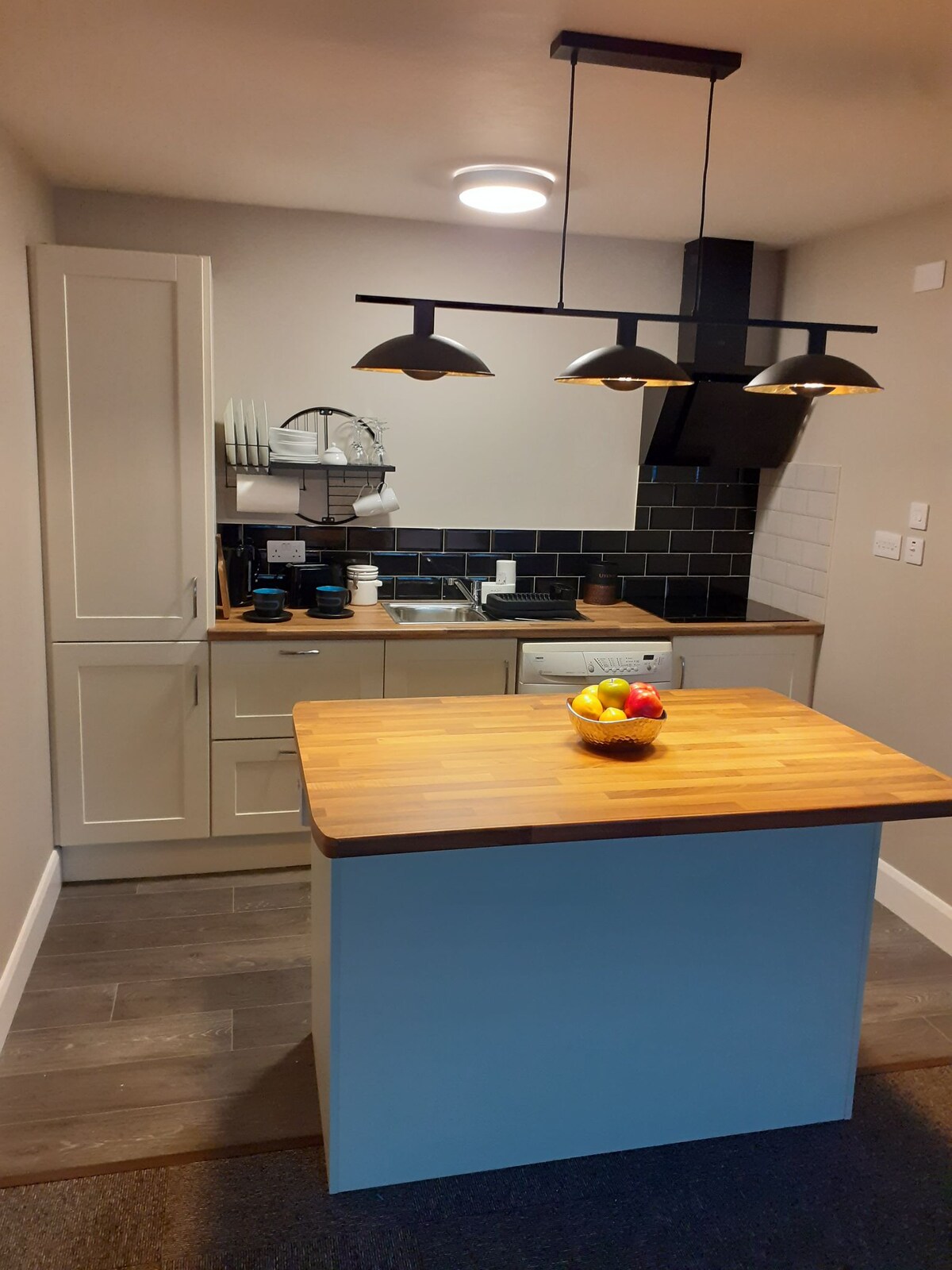 A modern kitchen area is featured, showcasing light cabinetry and a wooden countertop island with a fruit bowl. Pendant lights hang above the island, illuminating the space. A dark backsplash complements the kitchen appliances and accessories, creating a functional yet stylish atmosphere.