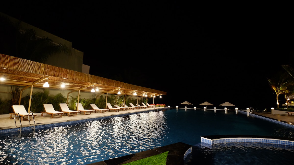 A spacious pool area is highlighted by soft lighting, creating a relaxed ambiance at night. Comfortable lounge chairs line the poolside, and umbrellas are visible in the background, complementing the tranquil water's shimmer.