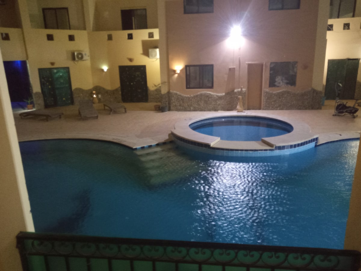 An inviting swimming pool is centered in a spacious courtyard, featuring a curved shape and textured stone borders. Surrounding areas are decorated with warm lighting, creating a serene ambiance, while lounge chairs are positioned for relaxation near the water's edge.