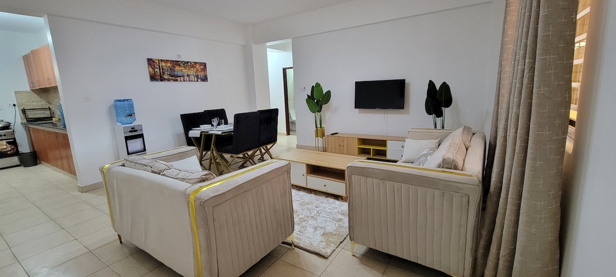 A modern living area is presented, featuring two light-colored sofas and a wooden coffee table. A flat-screen TV is mounted on the wall across from the seating arrangement. To the left, a dining table with four black chairs is visible, complemented by a well-equipped kitchen in the background.