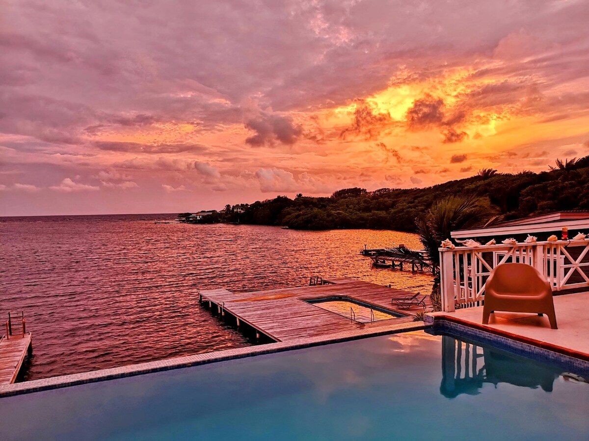 A serene view of the ocean at sunset is presented, with vibrant hues of orange and pink illuminating the sky. The infinity pool reflects the colorful sky, while a private dock extends into the water. Lush greenery borders the shoreline, enhancing the tranquil atmosphere.