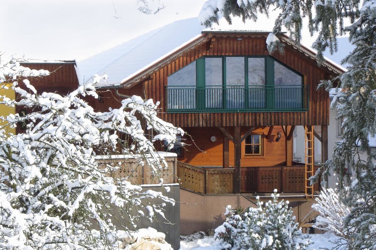A wooden house is surrounded by snow-covered trees, showcasing a large balcony with green shutters. The architecture features a steep roof and a warm wooden exterior, providing an inviting, rustic appearance against the winter landscape.
