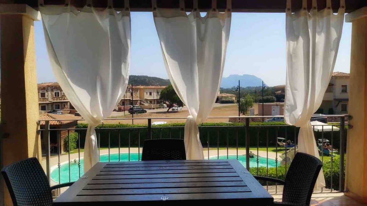 A shaded outdoor terrace features a wooden dining table and four chairs, overlooking a shared swimming pool. The view includes greenery and distant hills, with sheer white curtains gently flowing in the breeze.
