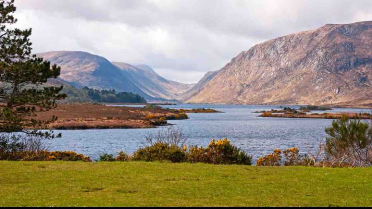 A tranquil landscape is depicted, featuring serene waters reflecting the surrounding mountains. Lush greenery frames the front of the image, leading towards the distant hills. The clouds above add depth to the scenery, creating a peaceful atmosphere that captures the natural beauty of the area.