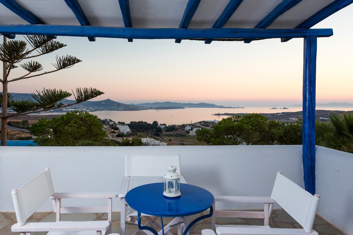 A private balcony features a round blue table surrounded by four white chairs. A lantern rests atop the table, with a backdrop of the serene sea view during twilight. Lush greenery is visible in the foreground, enhancing the tranquil setting.